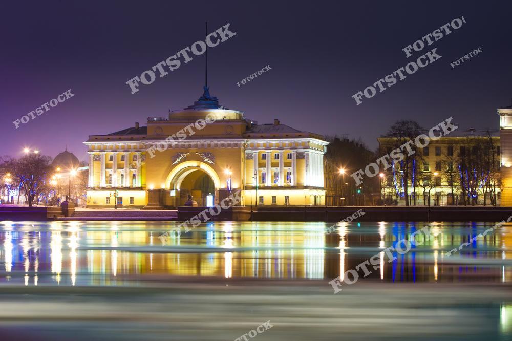 На снимке изображено здание Адмиралтейства, расположенное в центре Санкт-Петербурга, освещенное ночными огнями. Архитектурный ансамбль, выполненный в классическом стиле, выглядит величественно благодаря искусной подсветке. Перед зданием видна река Нева с красивыми отражениями света, создающими атмосферу уюта и тишины. Это одно из самых знаковых зданий города, связанное с его морской историей.